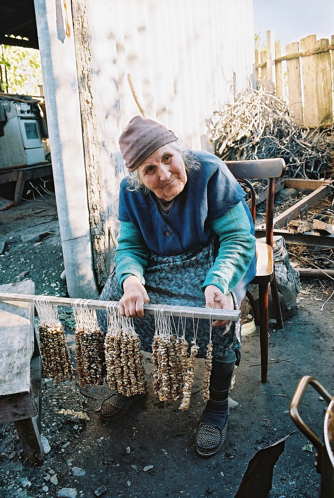 A seated woman holds a wooden rod hung with strings of nuts for making churchkhela.