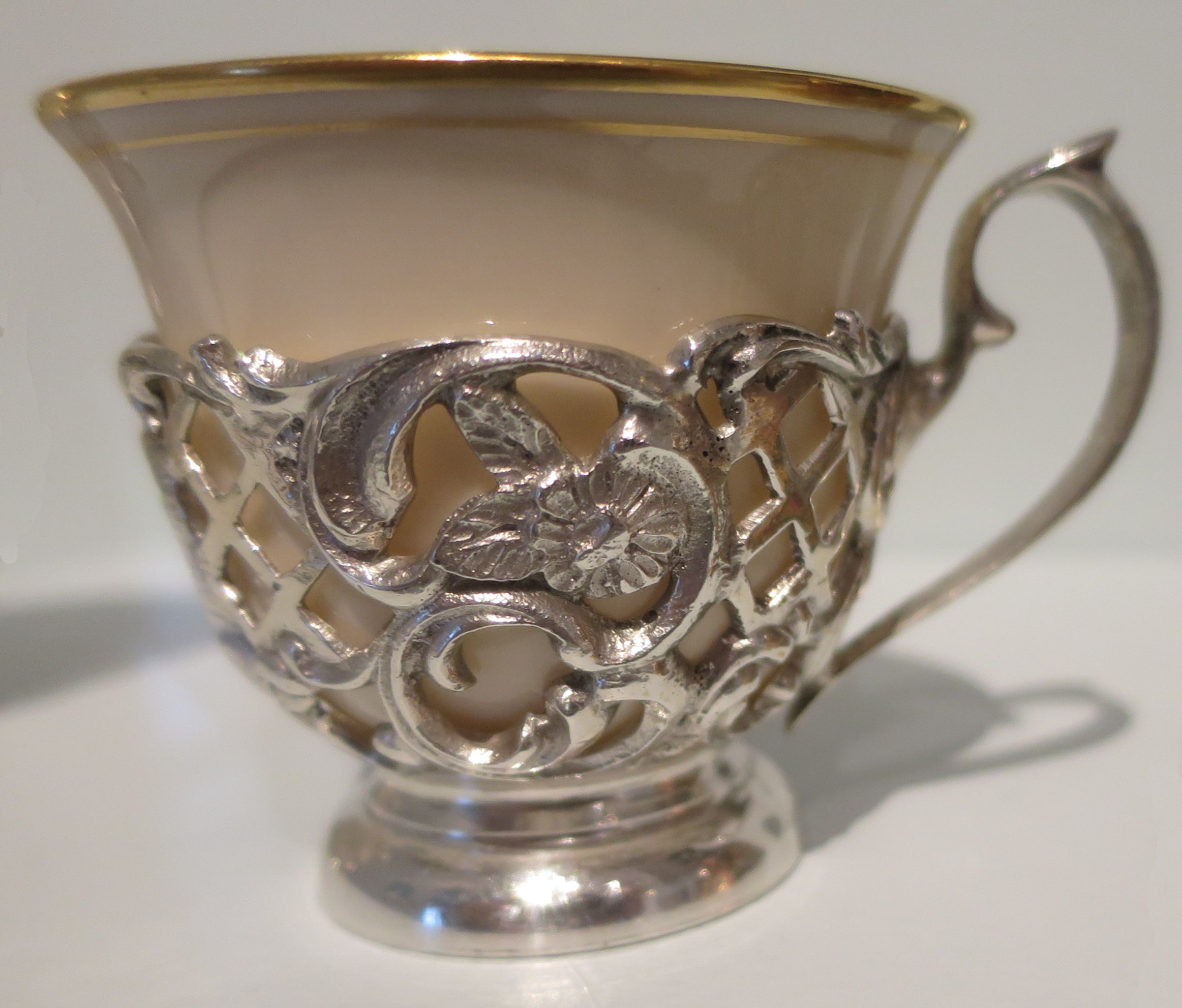 White porcelain coffee cup with a gold rim nested in an ornate openwork silver holder with a handle.