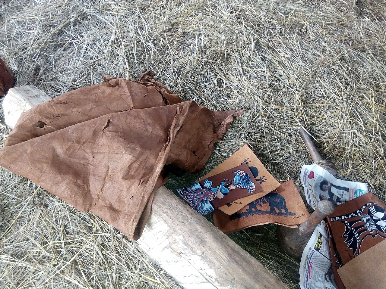 Brown barkcloth and decorated pieces displayed beside a log on dry grass.